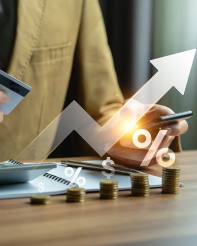 A business professional holding a credit card and smartphone, with a glowing upward arrow and financial icons, symbolizing growth, interest rates, and financial transactions. Stacked coins and a notebook are on the desk in the foreground.